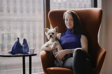 A beautiful young white European rich woman relaxed imposingly sitting in an armchair with a small Yorkshire Terrier dog on her lap. A girl with a pet at home. Vintage retro pale colors of interior.