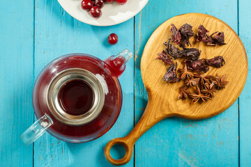 A teapot and a cup of hibiscus tea nearby on a blue table on a wooden board anise and cinnamon and hibiscus and cherry flowers in a plate. top view