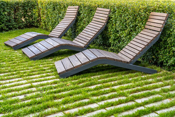 Three empty wooden chaise longues or deck chairs on tile and lawn among decorative bushes in...