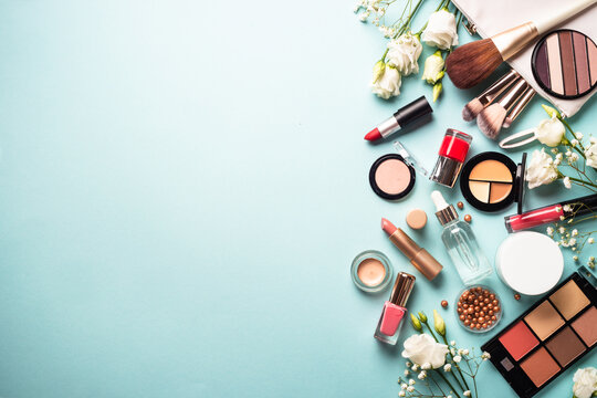 Cosmetic products on blue background. Cream, powder, shadow, brushes with green leaves and flowers. Flat lay image with copy space.