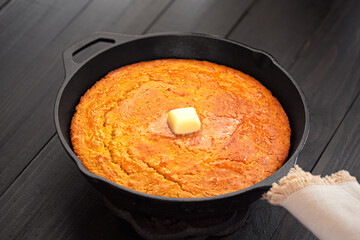 Freshly made corn bread in a cast iron skillet on a dark wooden surface with melting butter on top; copy space