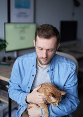 Caucasian male freelance software engineer sitting at working place in home office holding his ginger cat. Young developer man grey in t-shirt with domestic animal. High quality image