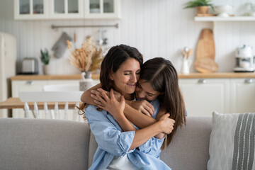 Happy smiling family of young mother and teenage daughter spending time together at home. Loving bored girl hugs woman sits on couch clinging to from behind feeling affection and not wanting to part - obrazy, fototapety, plakaty