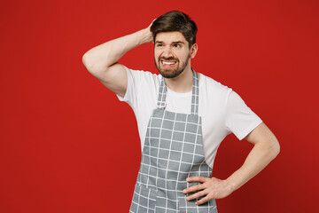 Young mistaken sad male housewife housekeeper chef cook baker man wearing grey apron look aside on area hold scratch head say oops isolated on plain red color background studio. Cooking food concept.