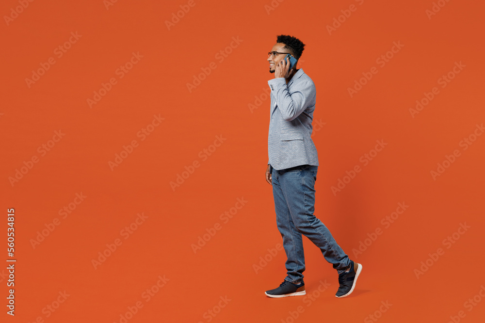 Wall mural Full body side view young employee business man corporate lawyer wear classic formal grey suit shirt glasses work in office talk speak mobile cell phone isolated on plain red orange background studio.