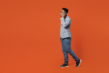 Naklejka premium Full body side view young employee business man corporate lawyer wear classic formal grey suit shirt glasses work in office talk speak mobile cell phone isolated on plain red orange background studio.