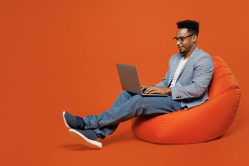 Full body young employee business man corporate lawyer wear formal grey suit shirt glasses work in office sit in bag chair hold use work on laptop pc computer isolated on plain red orange background.