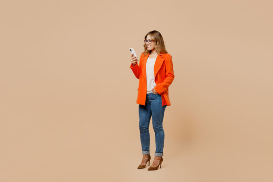 Full Body Young Happy Employee Business Woman Corporate Lawyer 30s Wear Classic Formal Orange Suit Glasses Work In Office Hold In Hand Use Mobile Cell Phone Isolated On Plain Beige Background Studio.