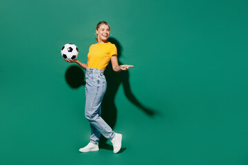 Naklejka na ściany i meble Full body fun young woman fan wear yellow t-shirt cheer up support football sport team hold in hand soccer ball watch tv live stream point index finger aside on area isolated on dark green background.