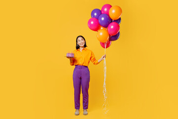 Full body happy fun young woman wears casual clothes celebrating holding in hand cake with candles look at bunch of balloons isolated on plain yellow background. Birthday 8 14 holiday party concept.