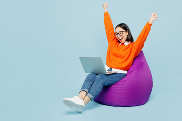 Full body happy young IT woman of Asian ethnicity wear orange sweater glasses sit in bag chair work hold use laptop pc computer do winner gesture isolated on plain pastel light blue cyan background.