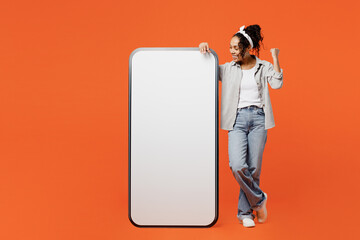 Full body young fun woman of African American ethnicity she wear grey shirt headband big huge blank screen mobile cell phone smartphone with area do winner gesture isolated on plain orange background