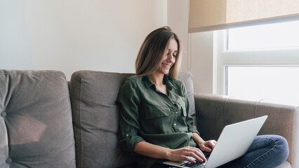 Young blonde woman using laptop at home. Beautiful caucasian girl smiling, sitting on sofa and working remotely with notebook. Noise and grain included.