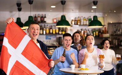 Screaming young adult sports fans rooting for favorite team and waving flag of Denmark while watching match together in pub