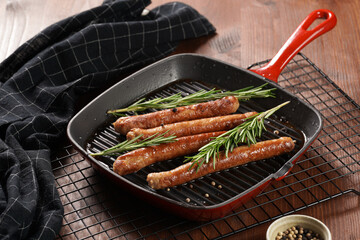 Traditional german organic pork sausages grilled on a red grill pan with rosemary branches and whole black pepper seeds on a baking rack on a wooden background
