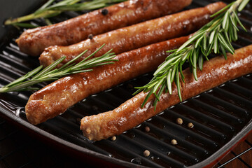Traditional german organic pork sausages grilled on a red grill pan with rosemary branches and whole black pepper seeds on a baking rack on a wooden background, close up