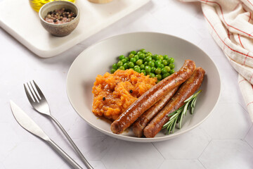 A lunch dish - boiled mashed sweet potato, steamed green peas and pork roasted sausages in a light grey bowl on a marble background