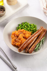 A lunch dish - boiled mashed sweet potato, steamed green peas and pork roasted sausages in a light grey bowl on a marble background
