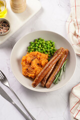 A lunch dish - boiled mashed sweet potato, steamed green peas and pork roasted sausages in a light grey bowl on a marble background