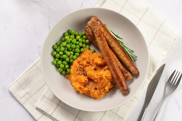 A lunch dish - boiled mashed sweet potato, steamed green peas and pork roasted sausages in a light grey bowl on a marble background, top view