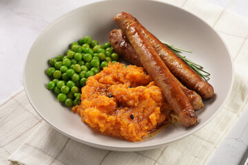 A lunch dish - boiled mashed sweet potato, steamed green peas and pork roasted sausages in a light grey bowl on a marble background