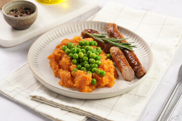 A lunch dish - boiled mashed sweet potato, steamed green peas and pork roasted sausages in a light grey bowl on a marble background