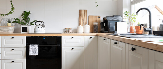 Cozy interior of kitchen with sink near window, wooden counter with kettle, oven and white kitchen furniture. Banner.