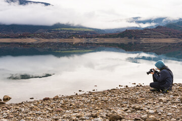 Teenager photographer taking picture to the reflection of the mountains with clouds in the calm water of the lake