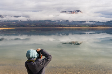 Rear view of teenage taking photos with his profesional camera to the refection of the cloud and mountain over the calm water of the lake