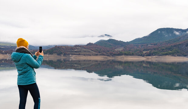 Hipster woman taking photos with her mobile phone of the amazing views of the mountain with fog reflected in the lake