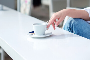 The hand of a girl with a manicure takes a cup with a drink. The girl takes a cup of coffee in her hand from a white table in a cafe on a summer food court. White cup of coffee on a white table