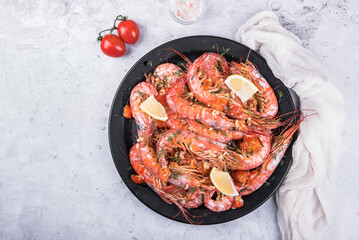 Baked langoustines in garlic sauce with cherry tomatoes on a black plate on a light marble background, top view. 