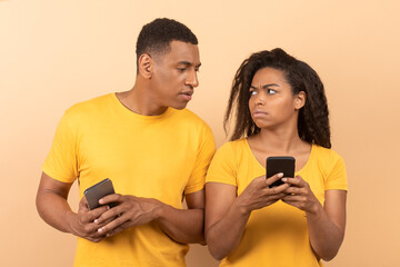 African american guy with smartphone spying on his wife chatting on mobile phone, yellow studio background