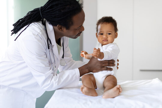 Smiling Pediatrician Doctor Talking To Black Infant Baby During Appointment In Clinic