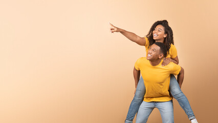 Excited black couple having fun, lady piggybacking guy and pointing at copy space, yellow...