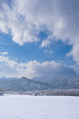 Fototapeta na wymiar 冬の晴れた日の雲の浮かぶ青空、山を背景にした雪に覆われた野と霜で白くなった木々。