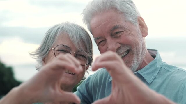 Close up portrait happy sincere middle aged elderly retired family couple making heart gesture with fingers, showing love or demonstrating sincere feelings together outdoors, looking at camera.