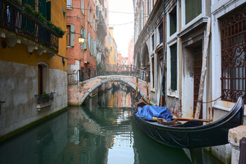 Beautiful view of canal with gondola parked next to the old walls, bridge decorated with flowers and picturesque old buildings surrounding the water under soft light in Venice, Italy.