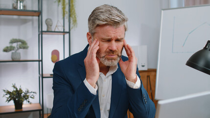 Tired ill mature businessman suffering from headache problem tension and migraine, stress at home office desk workplace. Middle aged freelancer broker worker man works on laptop notebook. Overworking