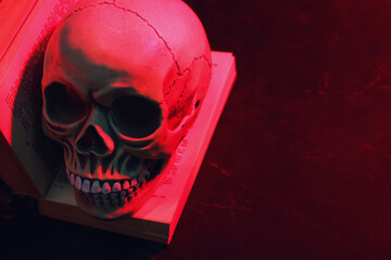 Human skull and old book in red neon light on table. Space for text