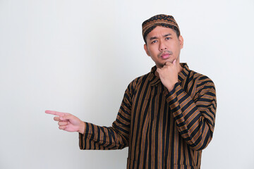 Asian man wearing traditional Javanese costume pointing to the right side with thinking expression