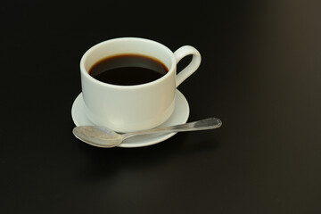 White porcelain cup of hot black coffee on a saucer with a spoon on a black background.