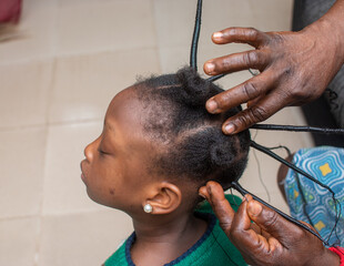 Naklejka premium African stylist making new hair style on the head of a little Nigerian girl child and rubbing cream on it