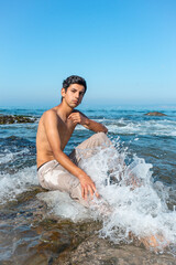 A dark-haired boy sits shirtless on the shore of the rocky beach with a wave crashing on his leg. Impressive water particles.