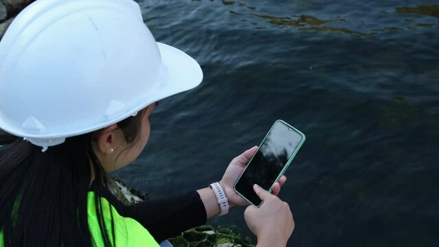 Female environmentalist using mobile phone to record water analysis data in dam. Researchers are collecting water samples from the river to test and detect pathogens. Water and ecology concept