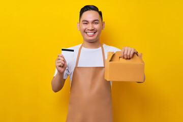 Smiling young Asian man barista barman employee wearing brown apron work in coffee shop holding paper takeaway cake bag and credit bank card on yellow background. Small business startup concept