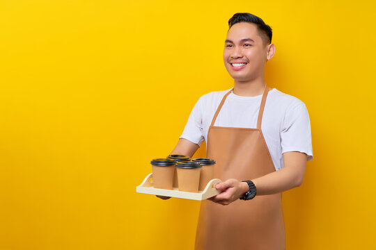 Smiling young Asian man barista barman employee wearing brown apron work in coffee shop giving takeaway paper cups of coffee or tea and looking aside on yellow background. Small business startup - Powered by Adobe