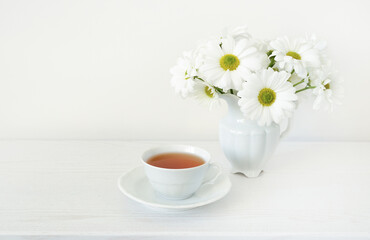 Bouquet of white flowers and white cup with tea on white wooden table, copy space. Herbal tea and bouquet of daisies or chamomile flowers