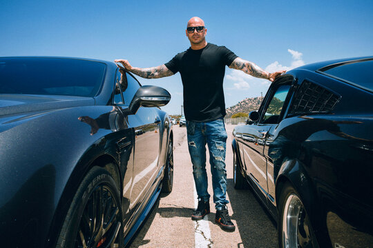 Man with shaved head standing in between two sports cars against a blue sky