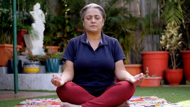 A middle-aged Indian housewife doing Pranayama or breathing exercises - morning routine. An elderly Indian lady sitting in a lotus position while practicing Yoga exercises - meditation  Yoga Asana ...